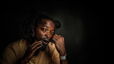 Ein Schwarzer Mann mit zwei Dutts und Bart berührt sanft sein Gesicht mit seinen Händen. Er trägt ein beiges Tshirt und eine Apple Watch. Sehr dunkles Farbfoto vor dunkelgrauem Hintergrund.