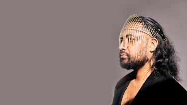 Ein bärtiger Mann of Colour mit schulterlangen lockigen Haaren trägt ein elegantes schwarzes Oberteil und futuristischen goldenen Schmuck auf der Stirn und dem Kopf. Er blickt nach links vor einem glatten lila-grauen Hintergrund.
